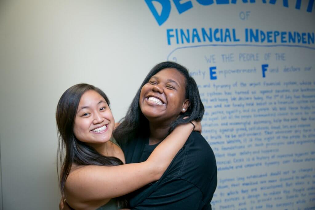 Two women hug and smile in front of a white wall that states the "Declaration of Financial Independence of the Community Empowerment Fund" in blue handwriting.